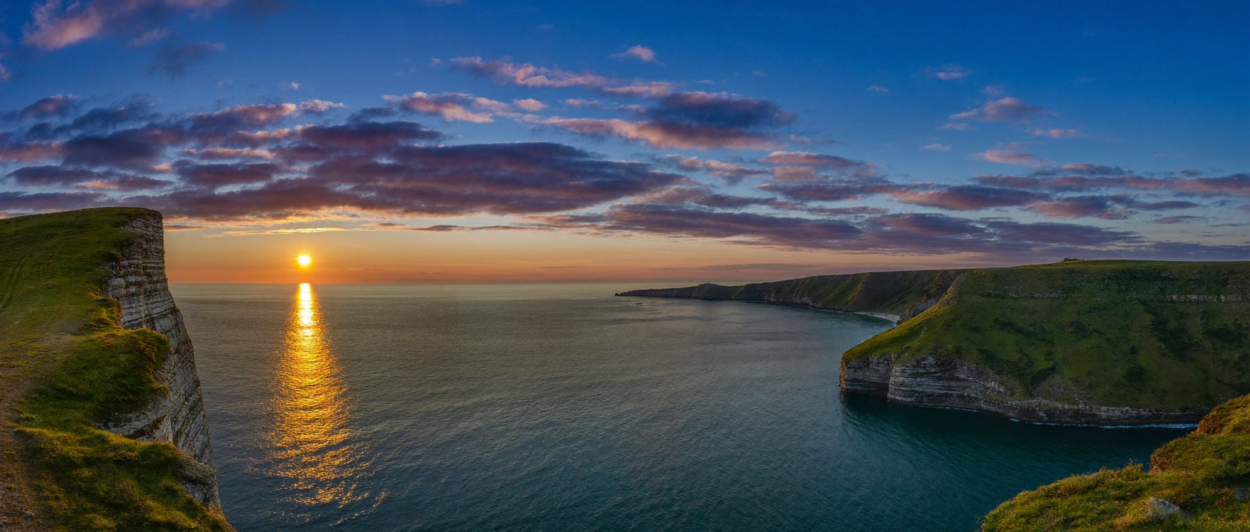 A wide clifftop view over the sea at sunset, green headlands framing the sun path