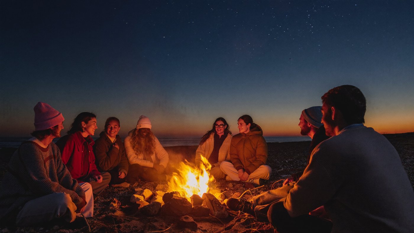 Friends around a driftwood beach bonfire at dusk under a deep blue twilight sky