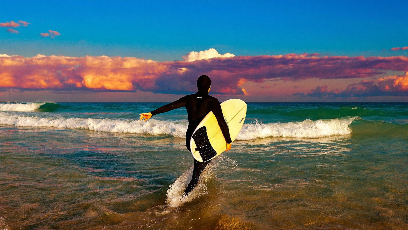 A surfer walking into the shallows at sunset, carrying a board
