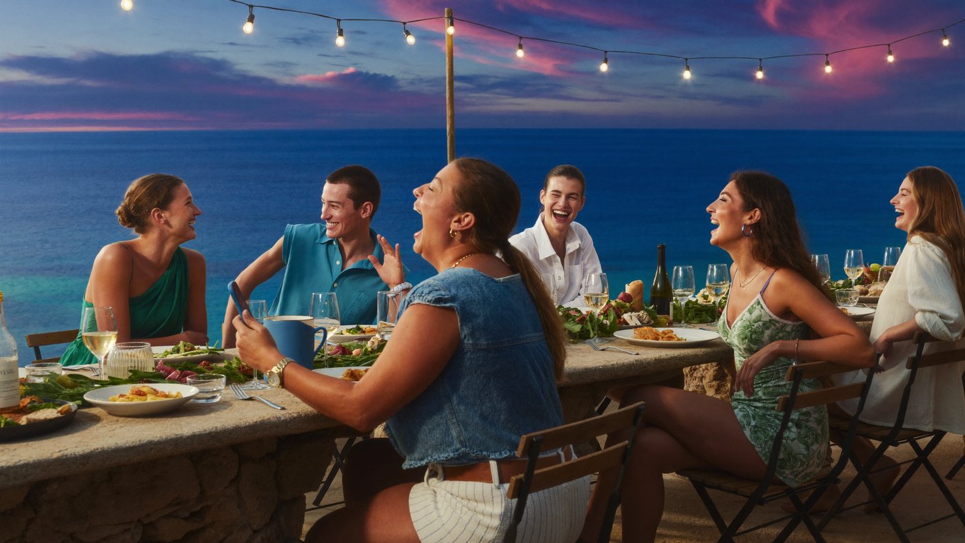 Friends at a long outdoor table above the sea at golden hour, nobody holding a phone